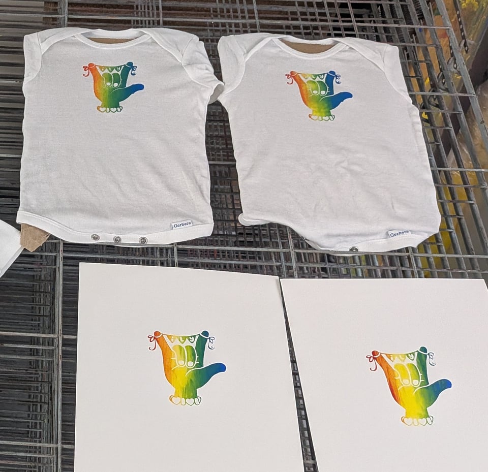 screenprints on a drying rack, two are white baby onesies and two are pieces of paper. The screenprinted image is a hand in the ASL handshape for I Love You, with a little banner stretched between the index and pinkie fingers that has the letters ILY on it. The screenprinted image is a rainbow gradient.