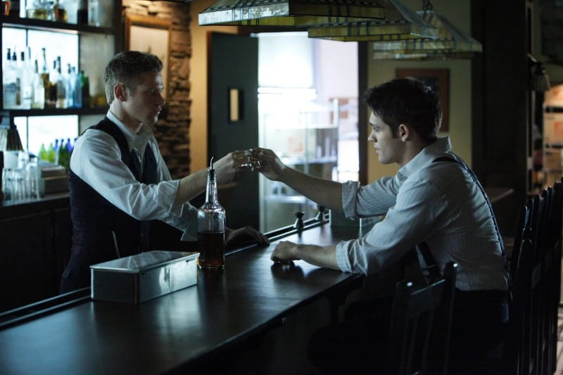 Two young men at a bar, one behind the counter and the other across from him on a stool, toasting with glass shots.