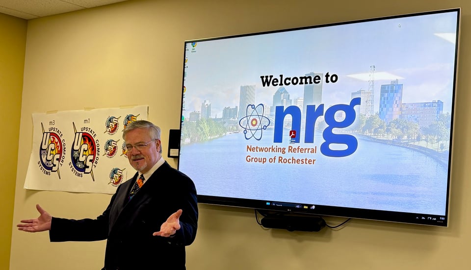 Darryl Powell gestures with open arms and hands facing up while standing in front of the NRG splash screen on a wall-mounted TV display.