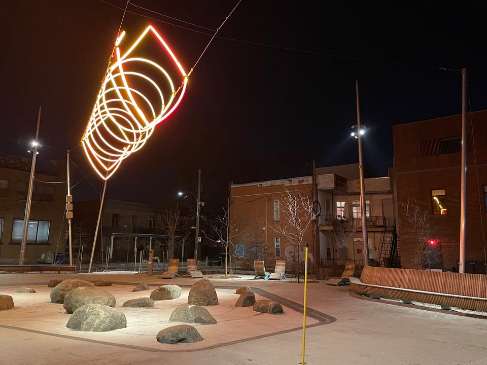 Image of a city park at night with benches, rocks and hard surfaces with suspended light sculpture hanging above it.