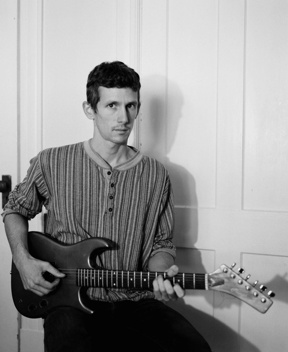 A man in a striped henley shirt holds an electric guitar as he looks at the camera.