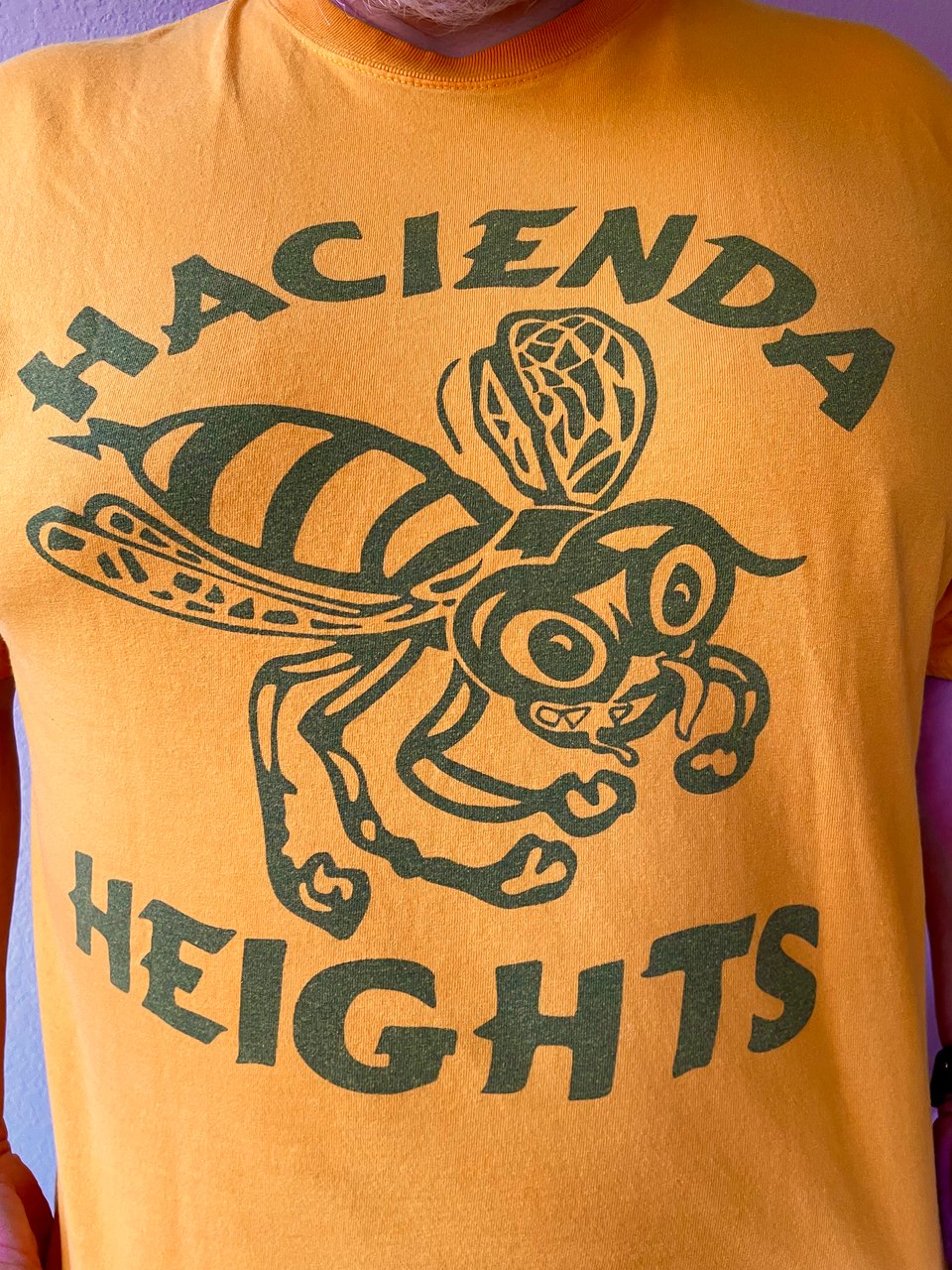 A man wears a yellow T-shirt with the words Hacienda Heights and a grimacing, cartoon Hornet on it. The logo and text are a dark green.