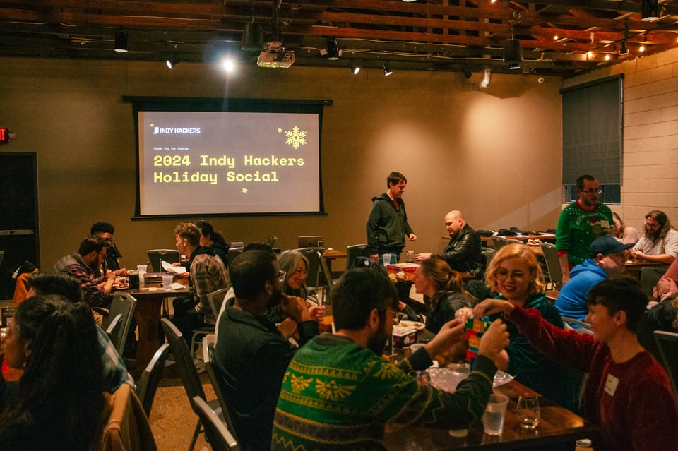 A projector screen with "2024 Indy hackers Holiday Social" displayed with an array of tables and event attendees playing games and spreading good cheer.