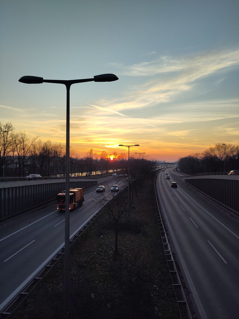 Blick auf eine mehrspurige, nicht zu dicht befahrene Autobahn. Am Horizont geht die Sonne unter, am Himmel ganz leichte Bewölkung.