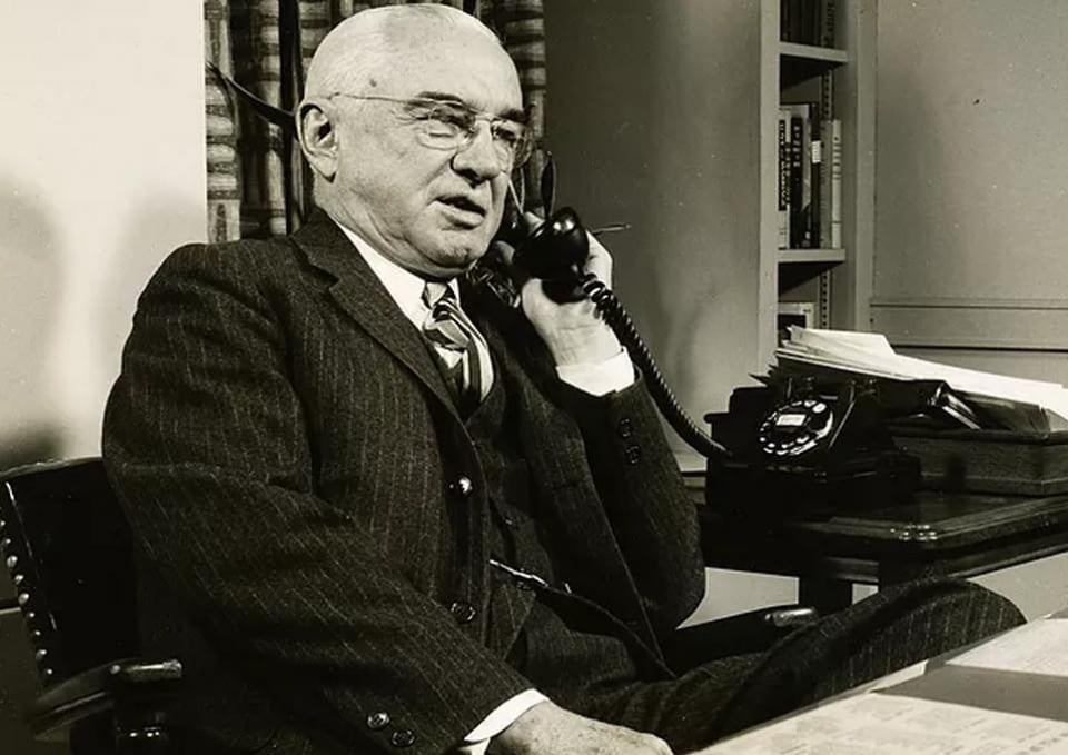 A black and white photo of an older white man with rimless glasses in a pinstriped suit on the phone. It's Alex Osborn.