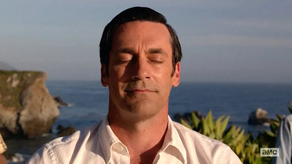 A man in a white shirt calmly meditates with eyes closed. Behind him, the ocean and cliffs are visible under a clear blue sky, conveying serenity.