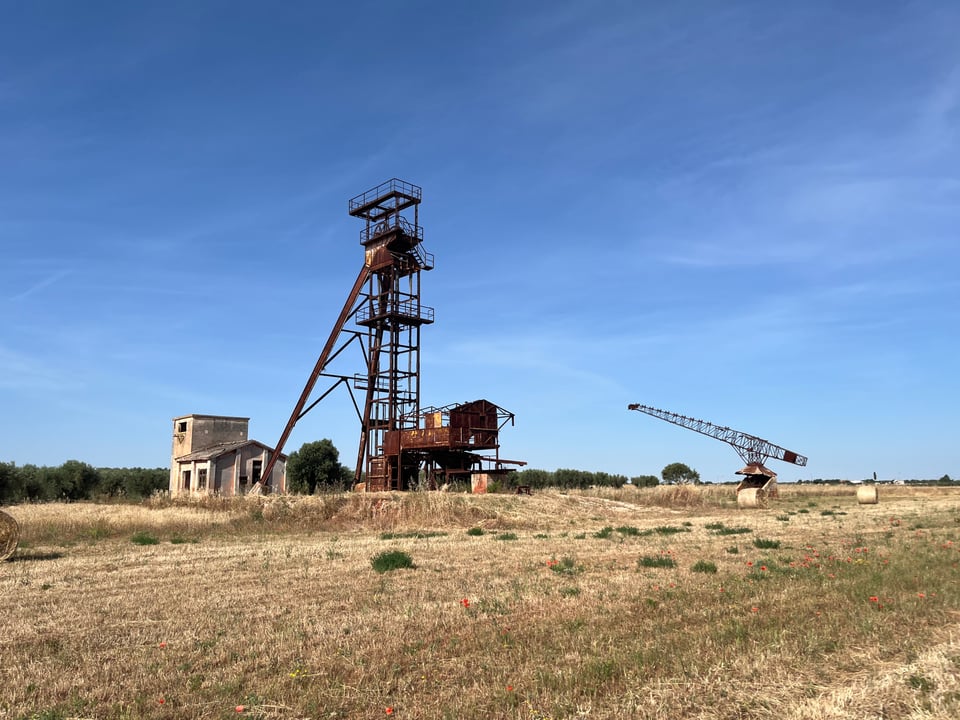 A tower and a crane, both brown rusted metal structures, against a bright blue sky, with a golden field in the foreground.