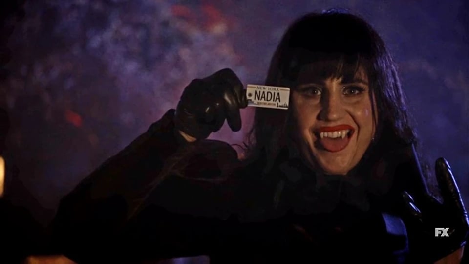 An enthusiastic Nadja from What We Do in the Shadows holding up a license plate that says NADIA