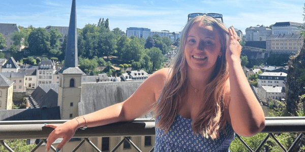 A JMC student with long blonde hair smiles while standing on a balcony, one hand touching her head and the other resting on the railing. Behind her, a scenic view of a European-style town with historic buildings, trees, and a prominent church spire is visible under clear blue skies.