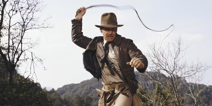 A picture of the actor Harrison Ford playing the character Indiana Jones, he is wearing a leather jacket, a beige shirt, and he is wielding a whip in the air. Behind are jagged hills and bare trees