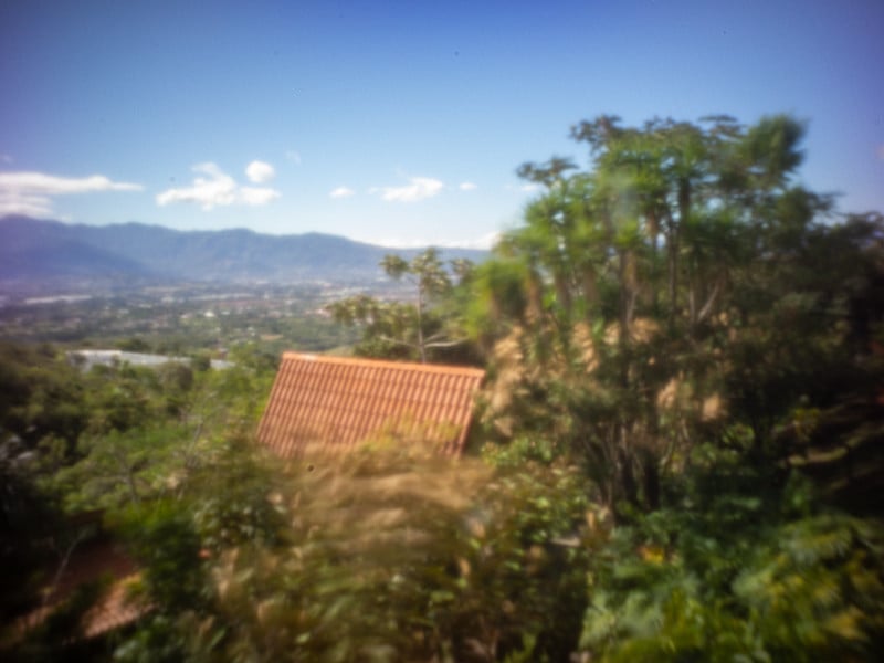 The roof of one of the lodging buildings, surrounded by lush vegetation, with mountains in the distance and a blue sky.  Everything is in soft focus from a pinhole camera