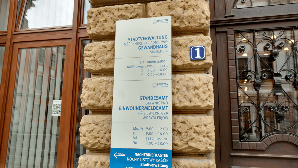 The sign outside a government office in Bautzen (Budysin), Germany, with the names of the offices and their opening hours. The text is in German and Sorbian. The German text is larger and in bold, and the Sorbian text is smaller and not bold and a little harder to read at a distance.