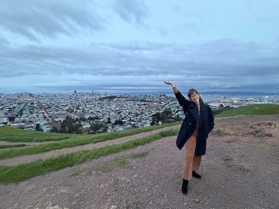 The author is wearing a dark blue peacoat and brown pants, smiling and gesturing up at the sky, on top of a hill in San Francisco. The hill is brown and rocky with green grass; downtown, the Mission district, and the bay are visible in the background; the sky is light gray with dark streaks.