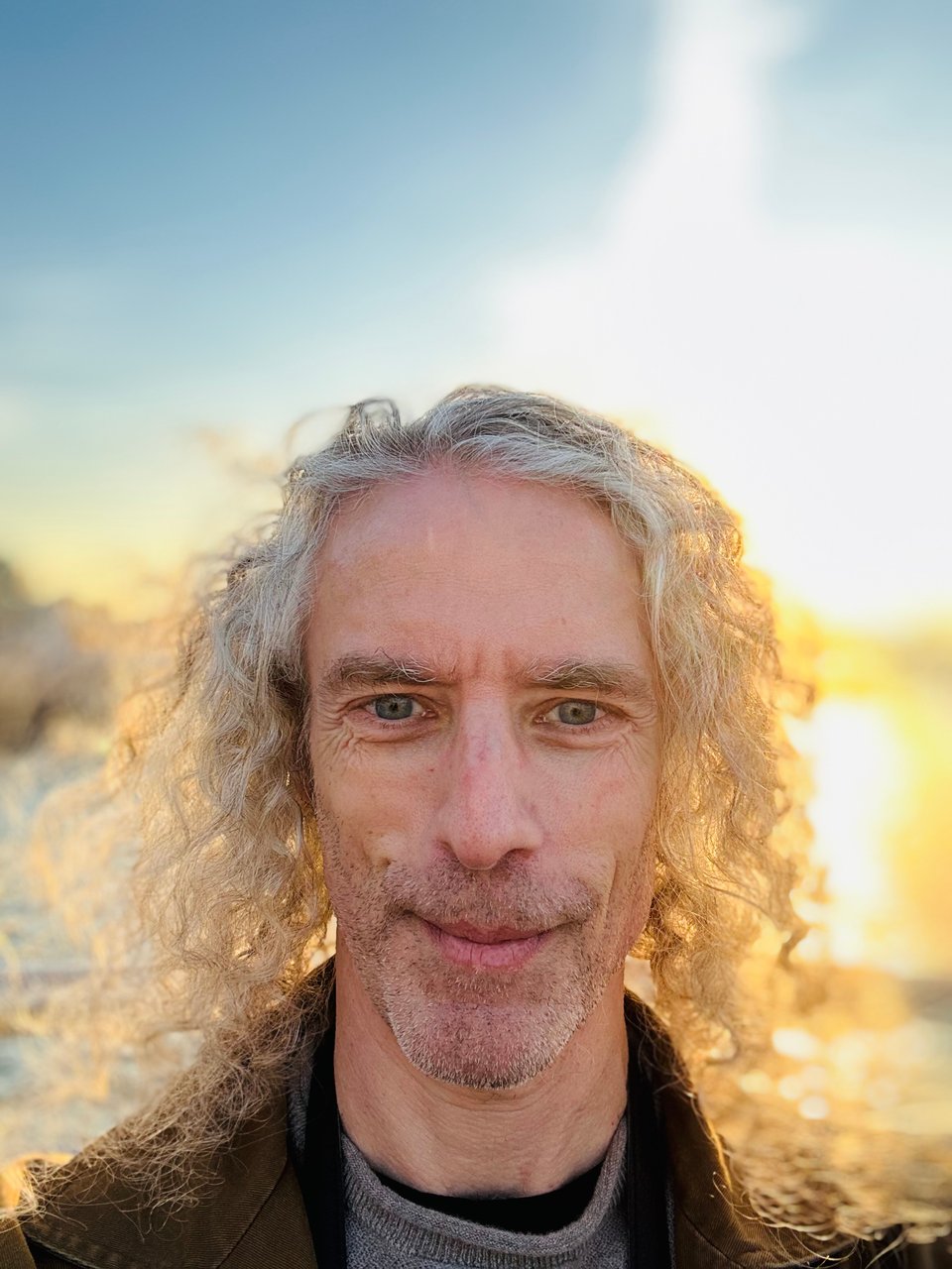 Selfie of white man with long curly hair, illuminated by low sun behind him on the Hudson