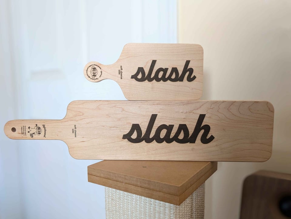 Wooden cheese boards with laser-engraved text that says ‘slash’ (as in slash fanfiction)
