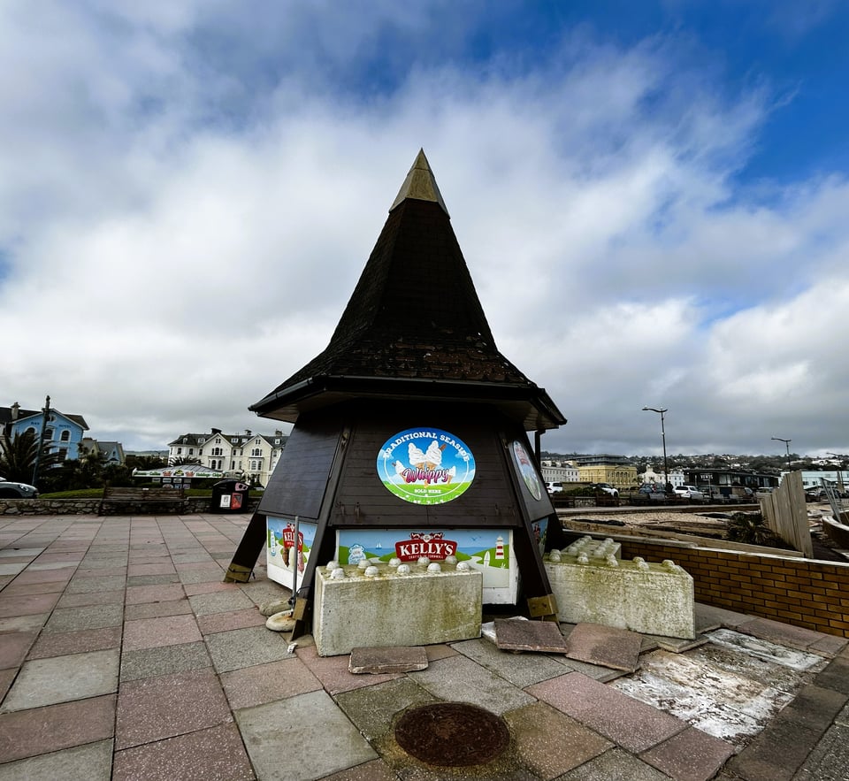 An ice cream hut. It's hexagonal with heavy wooden beams bolted to the ground and sloping up to a point. The serving hatches are shuttered for winter. Above them, the shingle roof looks like a pointy witch's hat. The patio beside it is yellow and pink paving slabs, some of which have been torn up. There are large concrete blocks with eight raised hemispheres on them - very much like a giant Lego brick - resting against the ice cream booth.