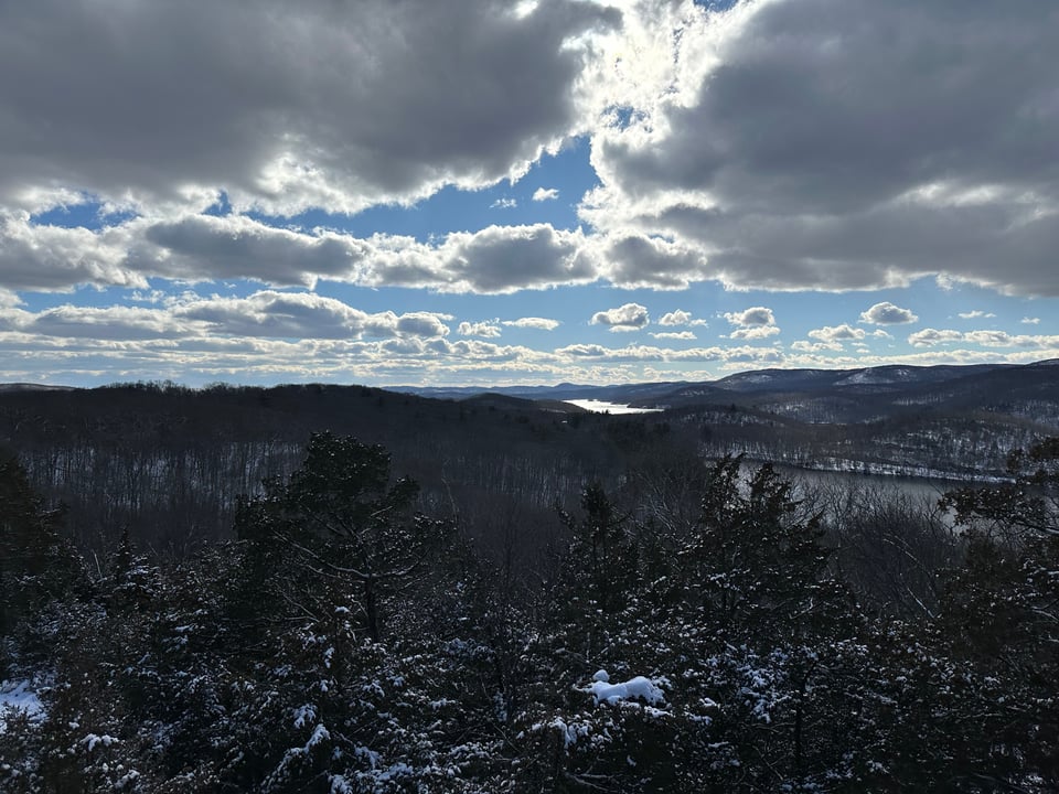 Photo of snow-covered woods, hills, and a reservoir, under partial clouds and a bright blue sky