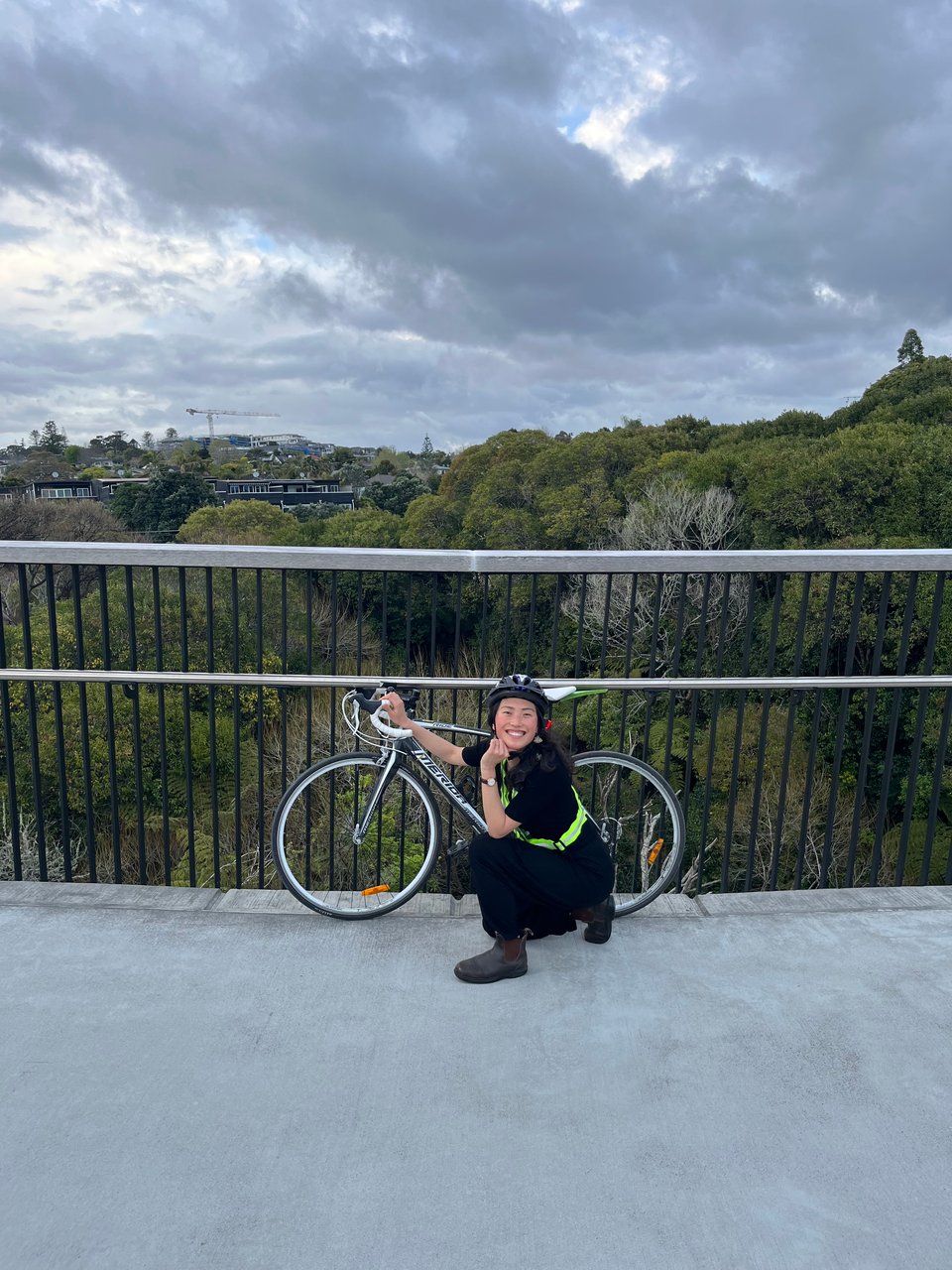 Photo of Jenny in front of a small white and grey road bike, on a smooth paved bridge with bush and city in the background