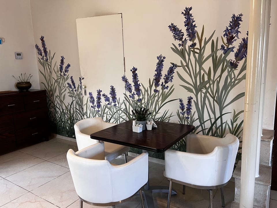 A hotel dining room with 3 white chairs around a dark wood tables. The wall is painted with a mural of lavender.  A centerpiece of lavender sits on the table