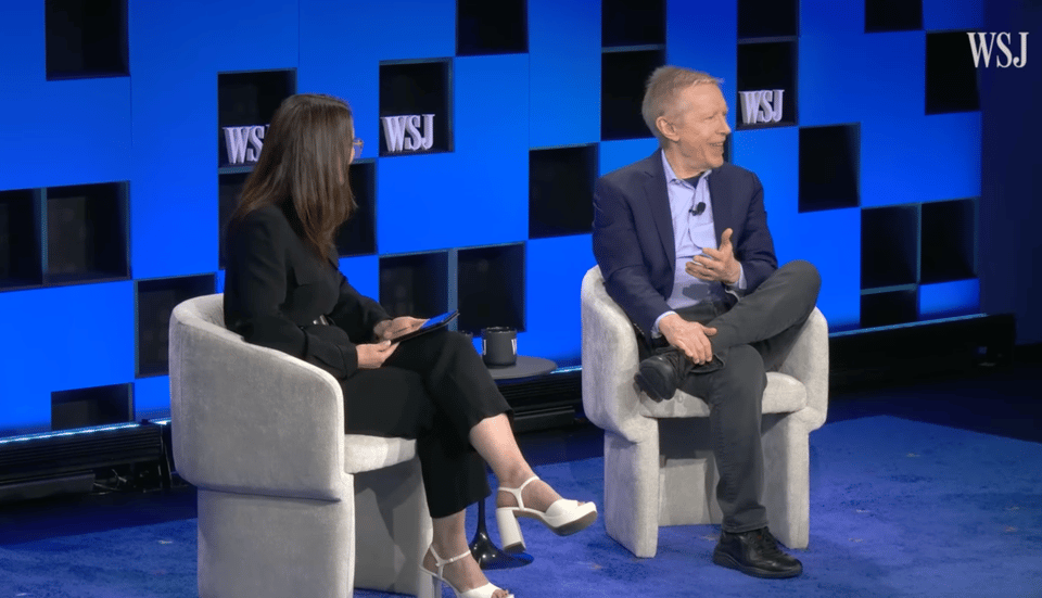 Two people sitting with crossed legs on a stage, with the "WSJ" logo on the wall behind them. The man on the right, Howe, is smiling at the audience and gesturing toward the woman on the right, Adamy, with his hand.