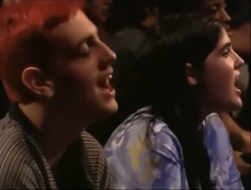 A still from a video of Alanis Morissette performing 'Thank U' in front of a live audience, showing two members of the audience singing along passionately