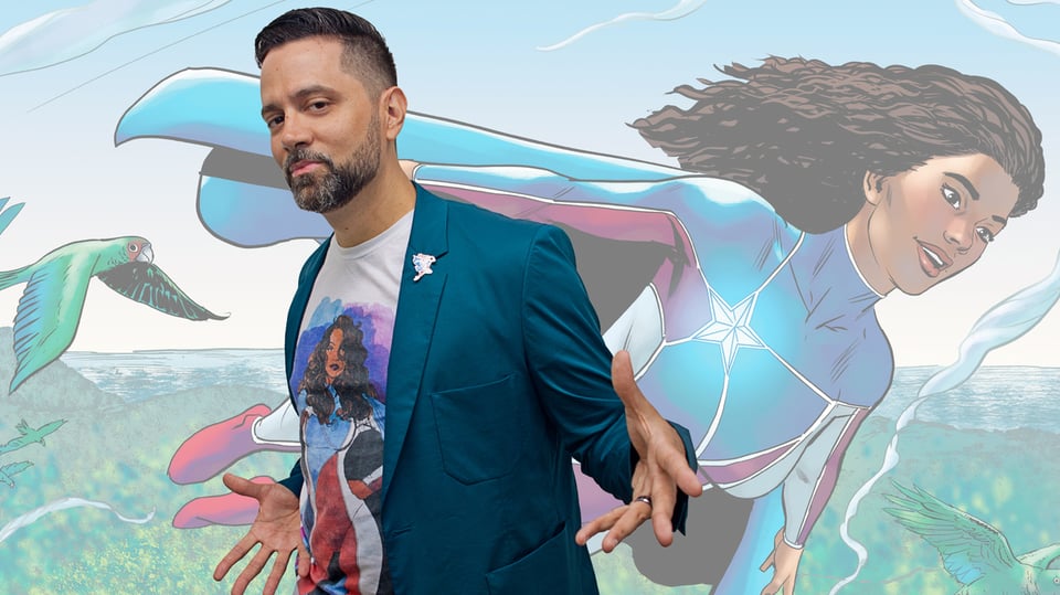 A Puerto Rican man with a high fade and beard in graying brown, wearing a tealish blazer over a T-shirt of his own creation and a white boutonniere, makes a presentative gesture over an image of La Borinqueña, the Puerto Rican superhero. She's a lady of ambiguous ethnicity, but obviously of color, who wears a costume based on the Puerto Rican flag: a blue cape and chest, with a white star over the right side, and red and white stripes along the arms and legs, ending in red boots.