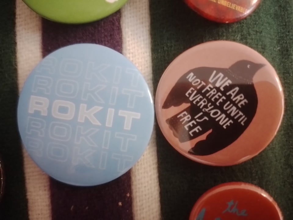 Two button badges. On the left, a pale blue one that says ROKIT in grey capitals. On the right, a salmon-coloured one with a drawing of a black bird, and the words We Are Not Free Until Everyone Is Free on the bird's back.