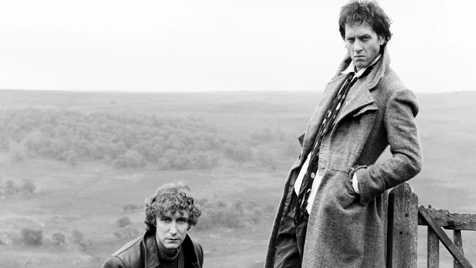 A moody, black and white photo of the two actors from the movie Withnail and I. On the left, crouching on the ground, is the actor Paul McGann, He has shaggy dark hair and is wearing a black leather jacket. Standing over him to the right is the actor Richard Grant, Wearing a thick tweed great coat with trousers, a white shirt, and a long necktie, and Grant is looking at the camera and scowling and leaning against a wooden fence. In the background is the countryside.