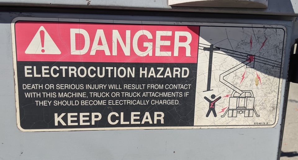 A sign that says DANGER ELECTROCUTION HAZARD.  Shows a cartoon figure standing next to a cherry-picker touching power lines. Red lightning bolts are striking the guy.