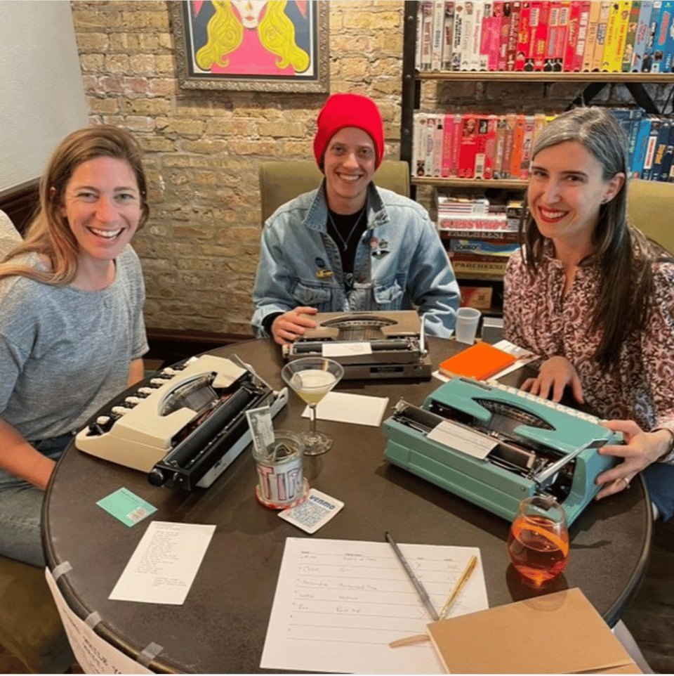 Three people are sitting at a round table, smiling at the camera. Each person has a vintage typewriter in front of them. There are also a couple cocktails, a sign-up sheet, and a tip jar on the table. Behind them, there is a brick wall with a framed movie poster and a rack of VHS cassettes organized by color.