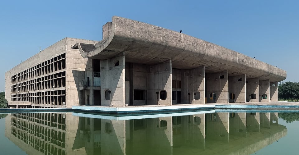 A large rigidly square building is fronted by a triple height entrance way made up of slim fin-like walls and a dramatically curved roof. The whole is reflected in a green pool.