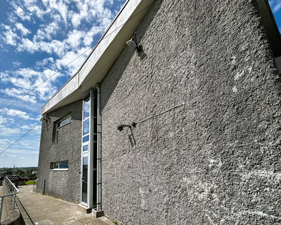 The side of the building. It's mostly roughcast concrete, with a single double height narrow window at the lowest point of the roof, and narrow strips of windows towards the front.