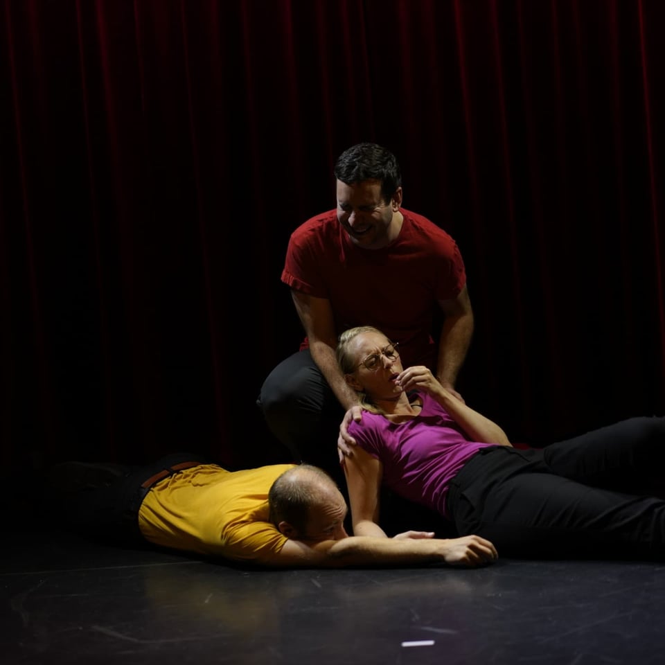 Three people on the floor, one laying down drinking something from a mimed class looking exhausted, one holding here, crying, and one laying on their stomach looking exhausted.