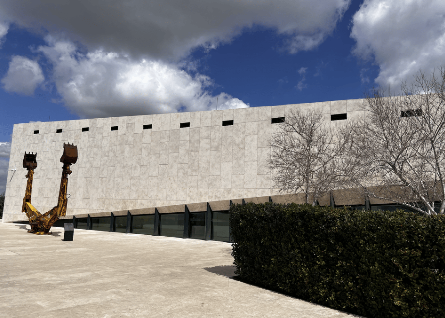 A light stone building against a blue sky with white clouds, with an art installation of the arms of two back hoes