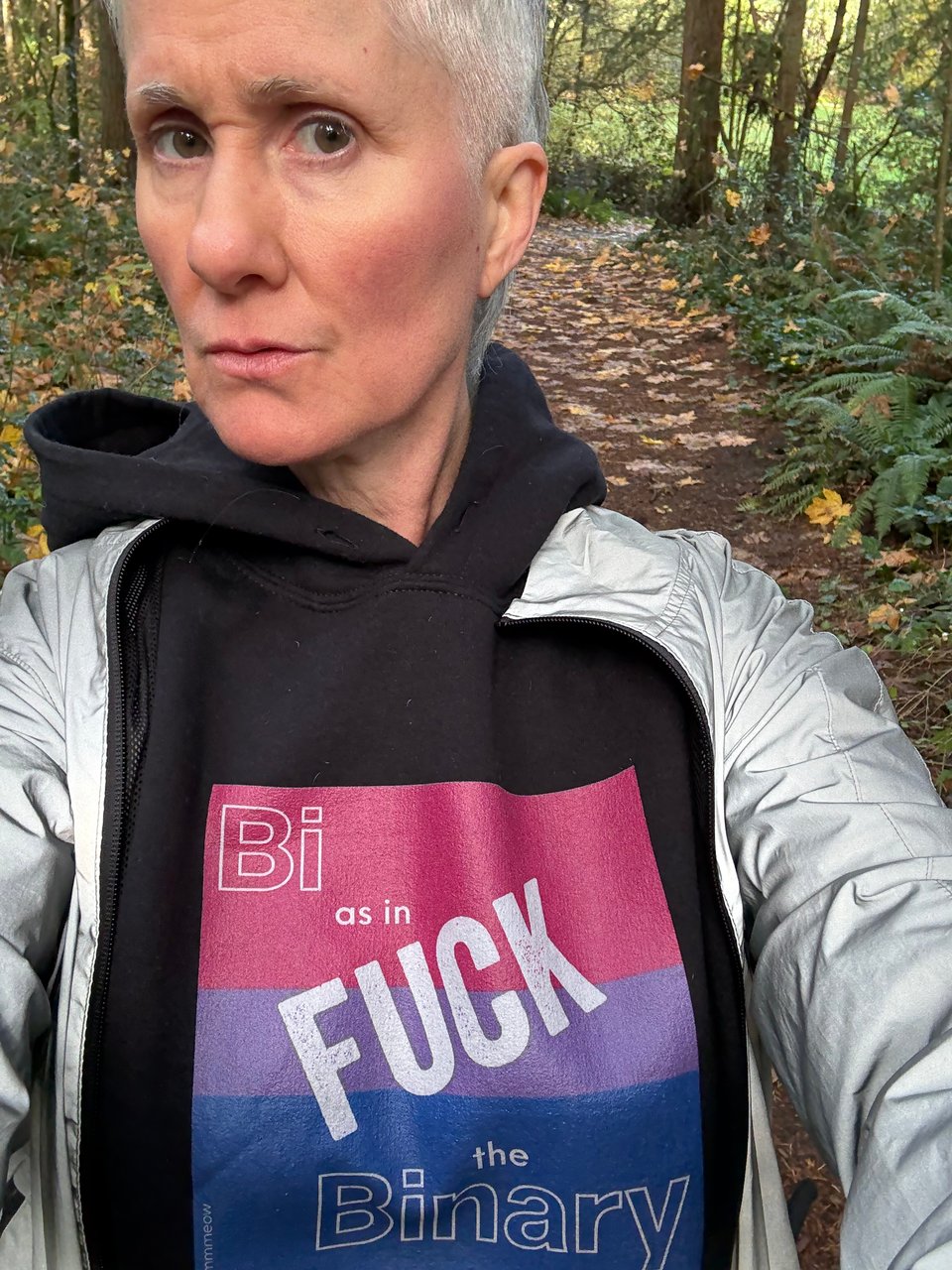 Me, a non-binary person with short grey hair, wearing a sweatshirt with the bi flag and the words Bi as in Fuck the Binary.