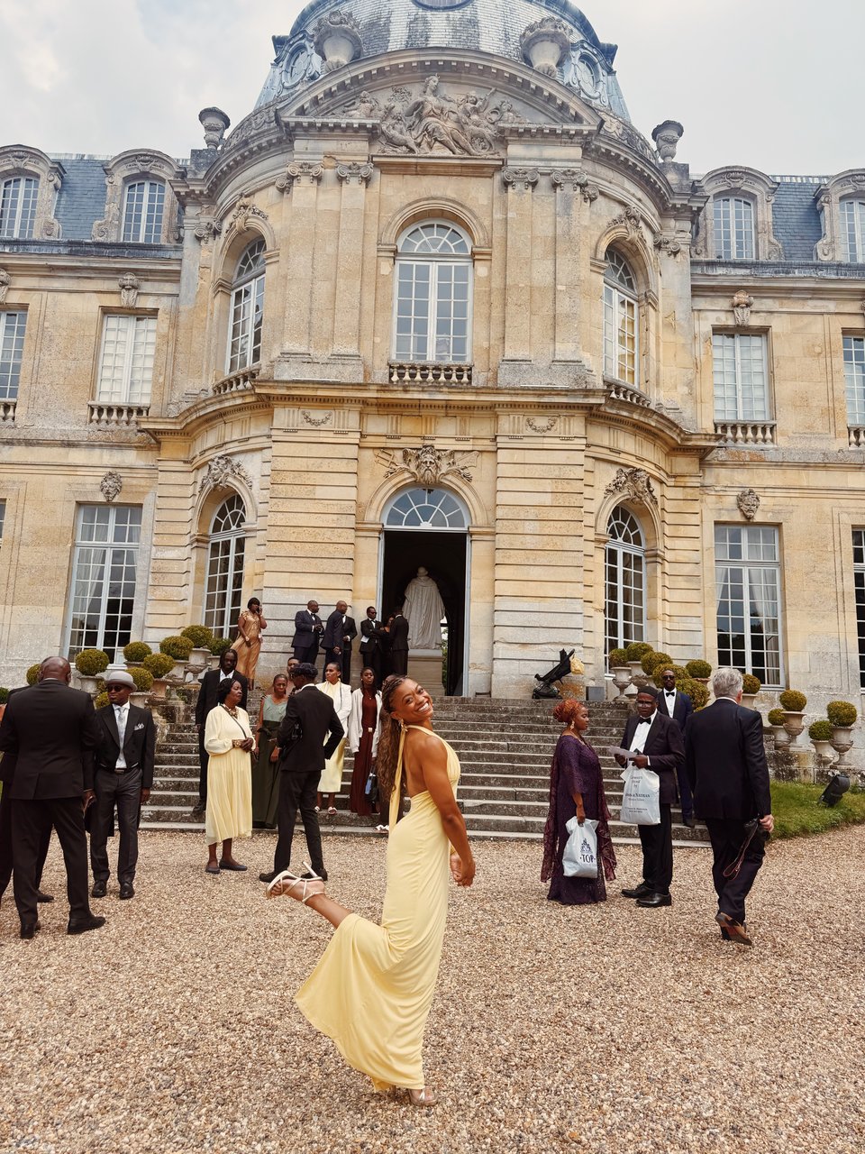 Nadia stands in front of a grand chateau, smiling brightly with one leg lifted in a playful pose. She wears a pale yellow halter-neck gown, strappy heels, and has pearls woven through her braids. Behind her, well-dressed guests gather on the stone steps and gravel path, with the elegant architecture of the chateau rising behind them.