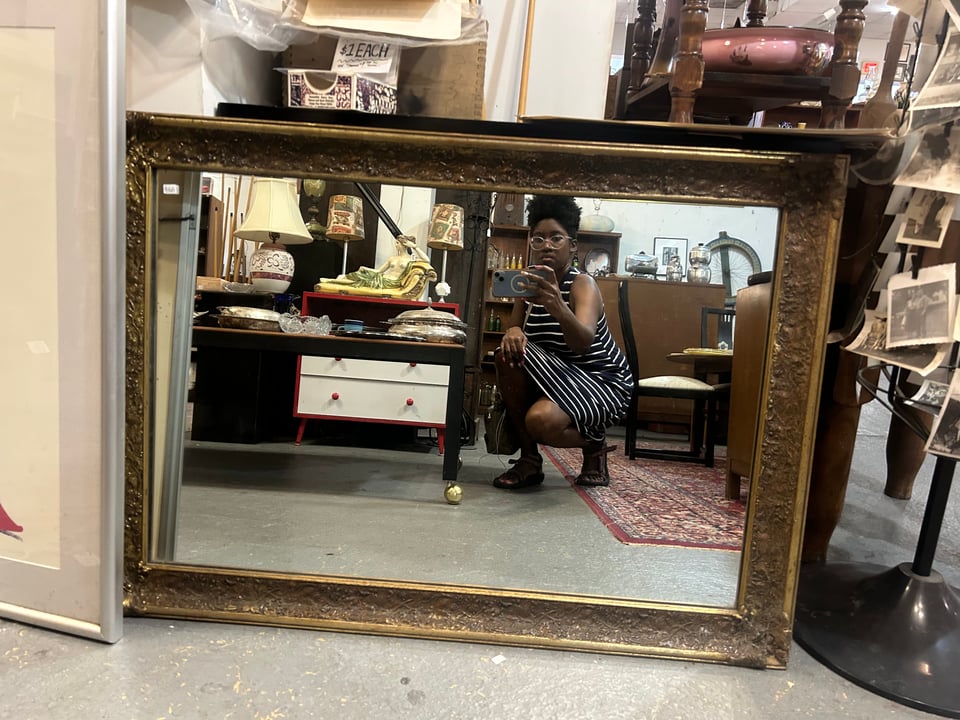 a photograph of a gilded mirror and a young, Black woman taking a picture with her phone in the mirror's reflection.