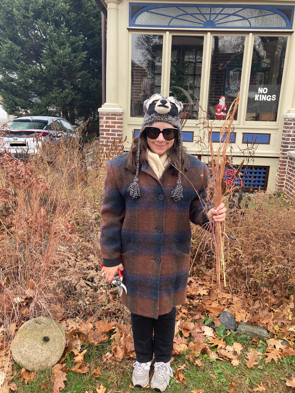 Woman in raccoon hat holding native plants in left hand in front of house. Native plant garden in background.
