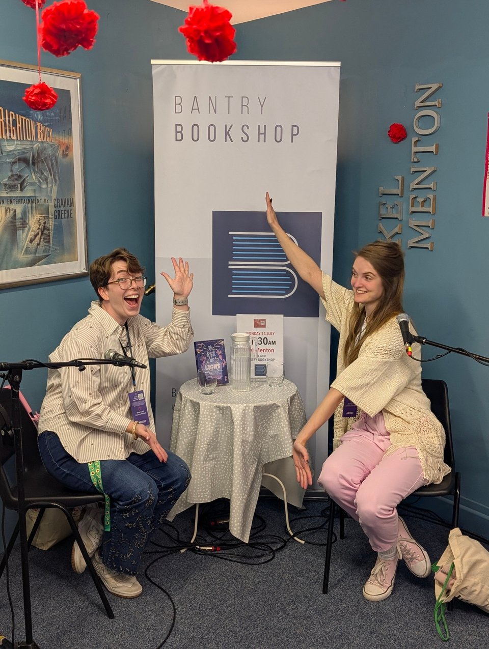 Kel Menton and Molly Twomey sit on either side of a small table in Bantry Bookshop. Molly gestures in a "ta da!" motion at Kel. Kel does the same back, their arms are not spread as wide.