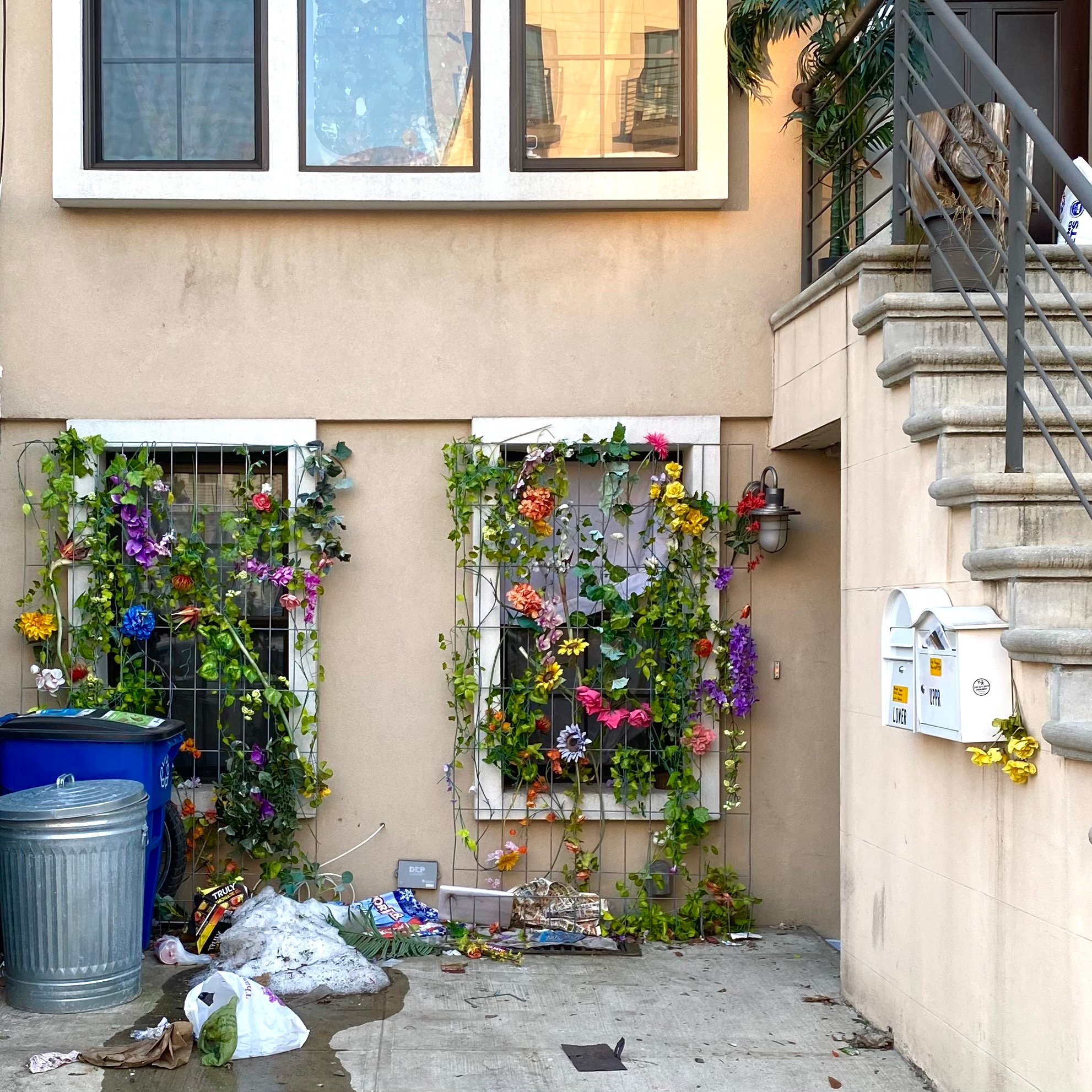 Image of a front yard of a white stucco house, the lowest floor of which has two windows with grids of purple, pink, yellow, blue, orange, and red flowers with green vines hanging all over it. The flowers may be fake but the trash in front of the house is definitely not, as they are scattered all over the place in front of what is otherwise a pristine home. To the right, there is a stone stairwell leading up to the brown door, with two white mailboxes attached to its left side.