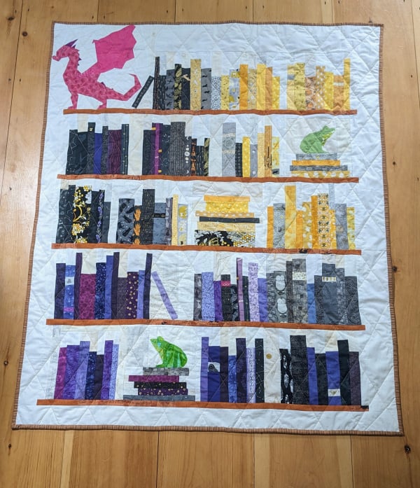 A lap quilt with five shelves of books arranged in a nonbinary yellow-to-purple gradient, black and grey at the edges. Simplified frogs sit on some shelves and a big pink dragon in the upper left.