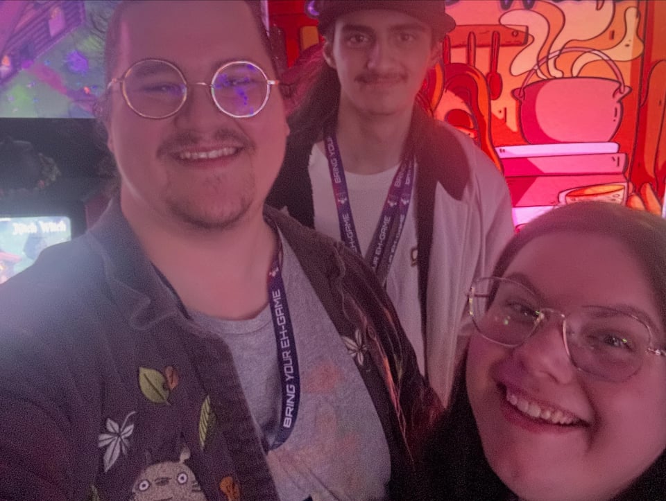 A selfie-style shot of three teammembers from Mistik Studios. Behind them is a screen, a banner of their game, and glowing pink light.