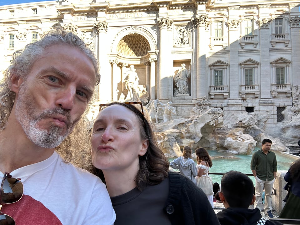 Selfie-photo of a white man & woman making kissy-faces at the camera in front of Trevi Fountain in Rome.