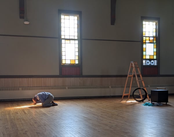 Tall windows in a high-ceilinged room cast sun puddles on worn wood floors through golden stained glass. I am kneeling, rocking forward, in the left-hand sunbeam, hands to my face, in business clothes. In the right-hand sunlight is a set assembly of ladder, box, wheelchair wheel.