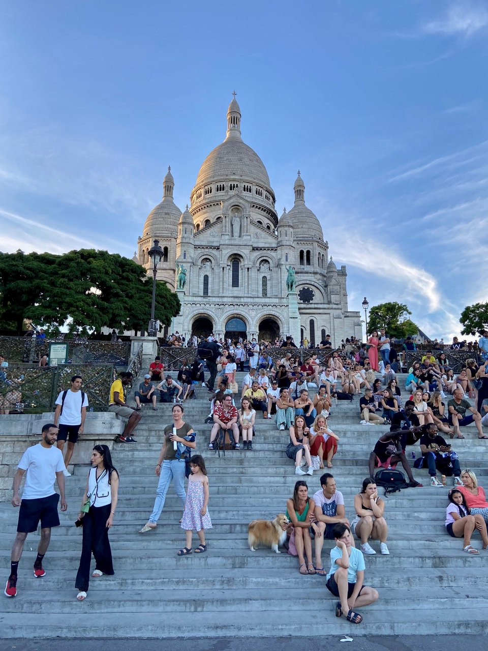 Photo of the steps of Sacre Cour in Paris, France, with many people (and a Corgi) sitting on the steps, with the cathedral in the background under a gorgeous blue sky