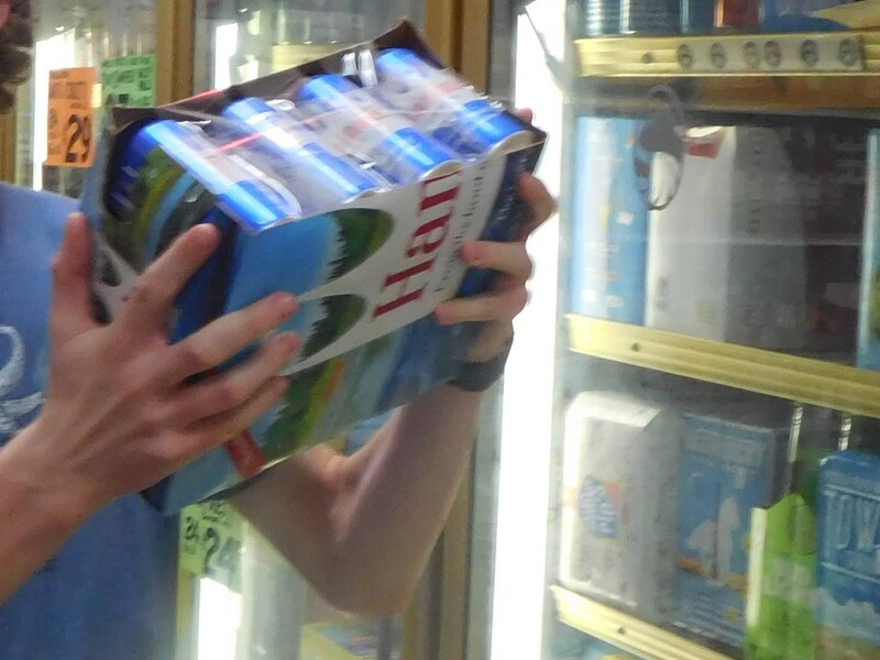 Two hands hold a case of Hamm's beer in front of a liquor store's chilled shelves