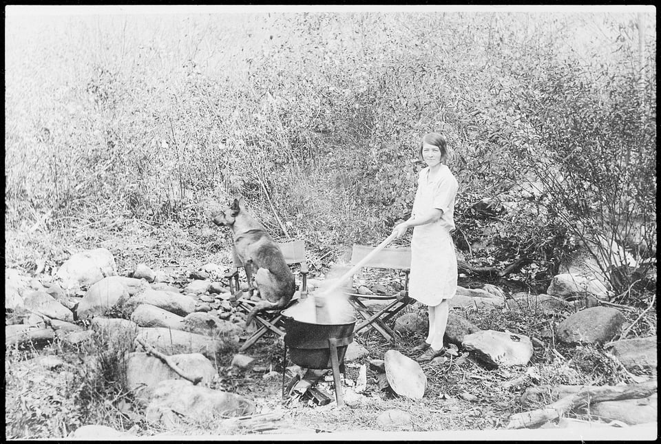 A woman uses a long stick to stir a steaming pot of apple butter over an outdoor fire. A German shepherd sits on one of two camp chairs.