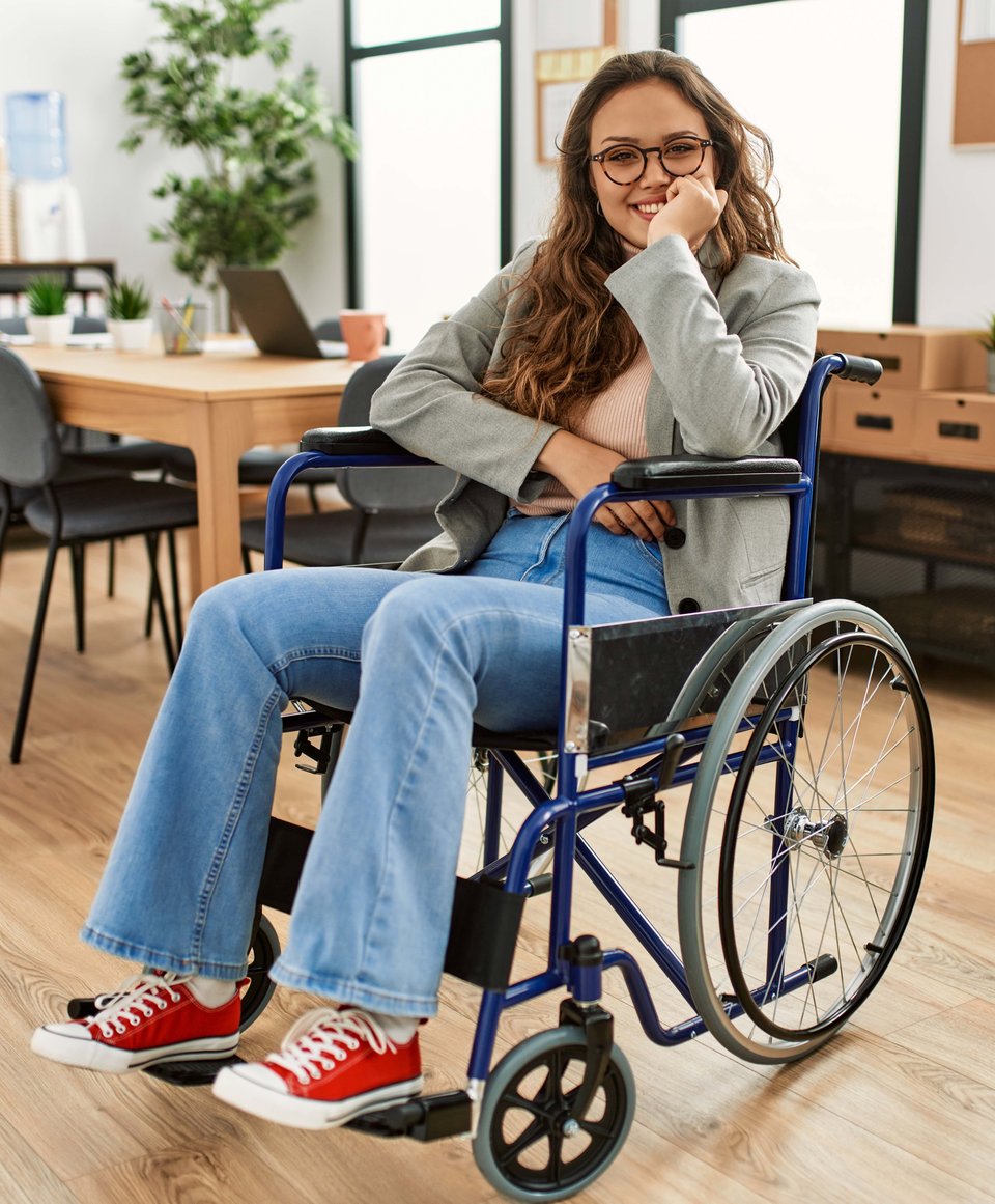 A beautiful woman with glasses sits in a blue wheelchair in an office. She's smiling with her arm rested on the arm of her chair, and cradling her face. She's wearing red Converse sneakers.