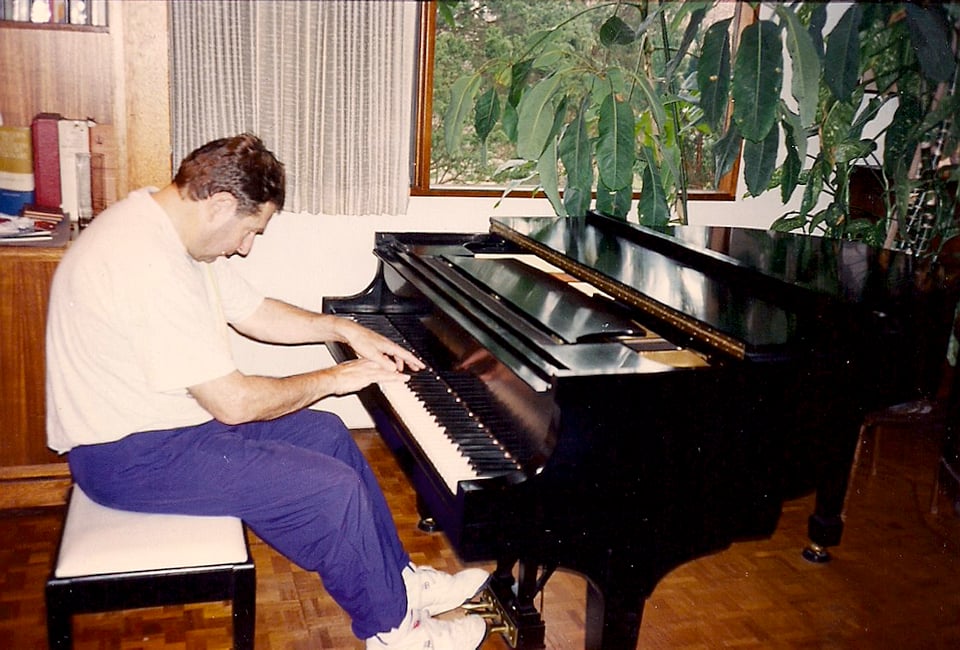 Man in white tee shirt and purple sweat pants playing Steinway baby grand piano next to large potted house plant