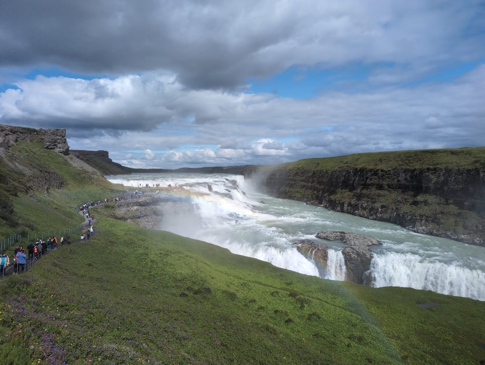 A photograph of Gullfoss, a huge, multitiered waterfall in Iceland. A line of people snakes along the path toward it on the left; in the foreground is a rainbow caused by the mist.